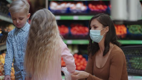 A-mother-in-a-protective-mask-with-two-children-is-buying-groceries-at-the-supermarket.-Buying-food-vegetables-and-fruits-with-children